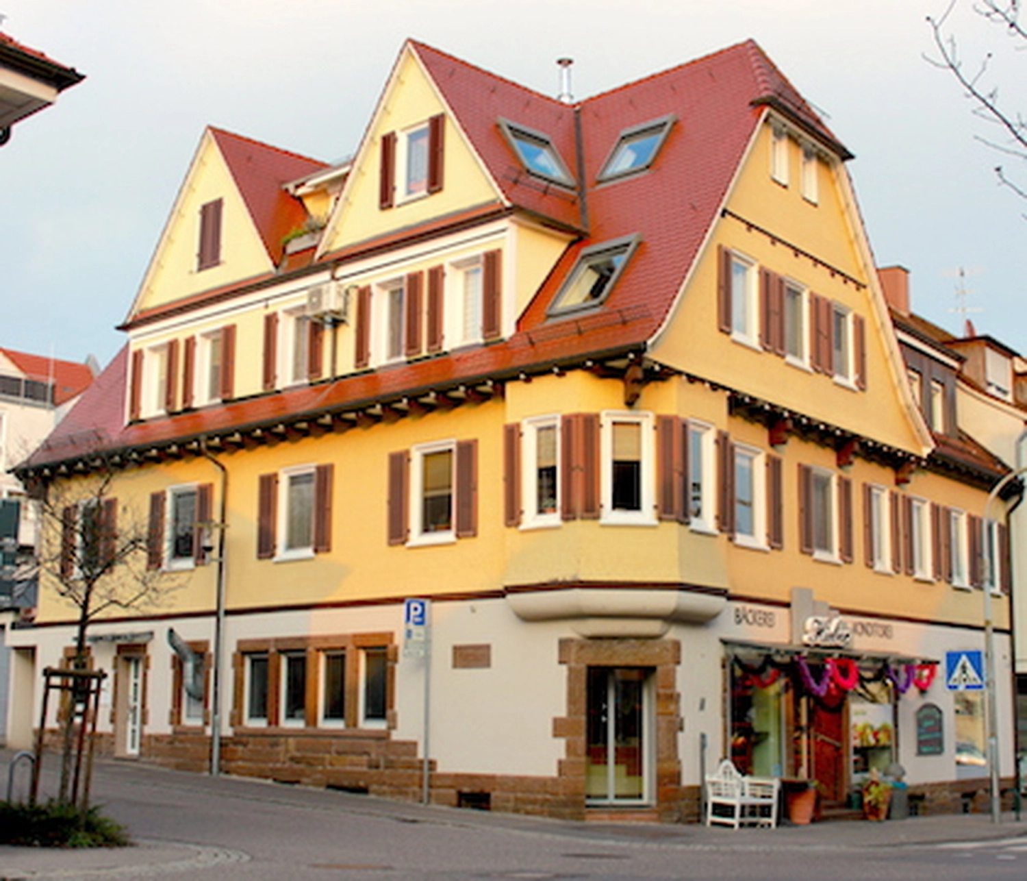 Hieber GmbH – Bäckerei – ein Eckgebäude im traditionellen europäischen Baustil mit gelber Fassade, rotbraunen Fensterläden und rotem Ziegeldach, das im Erdgeschoss einen kleinen Laden oder eine Bäckerei beherbergt.